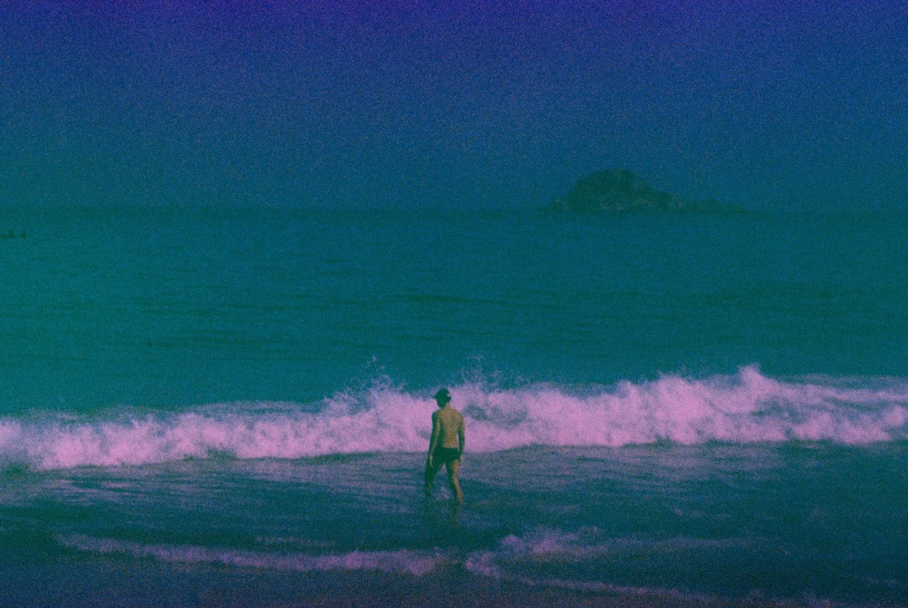 Foto do editorial: pessoa à distância entrando no mar, montanha ao fundo.